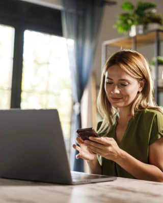 Frau sitzt zu Hause am Tisch mit offenem Laptop und Smartphone in der Hand